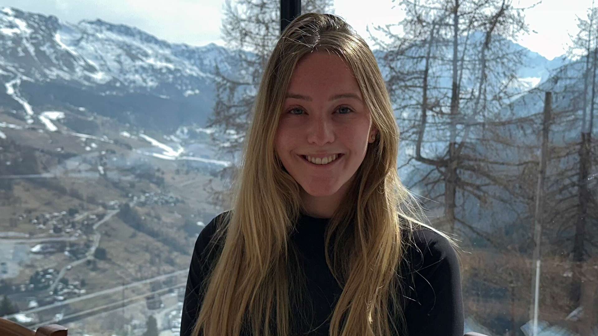 A young woman with long blonde hair standing in front of a mountain.